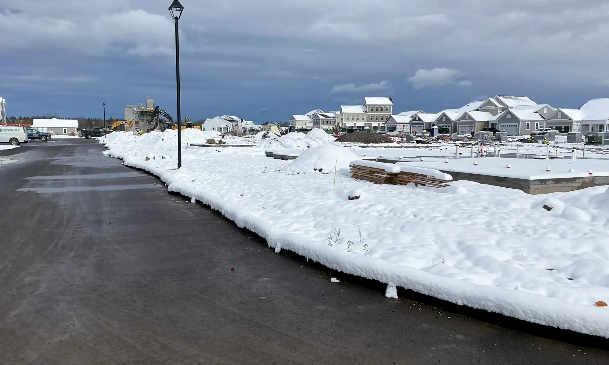 Active residential construction site during winter showing snow-covered building materials, equipment, and partially completed homes under overcast sky at Arbors at Penfield
