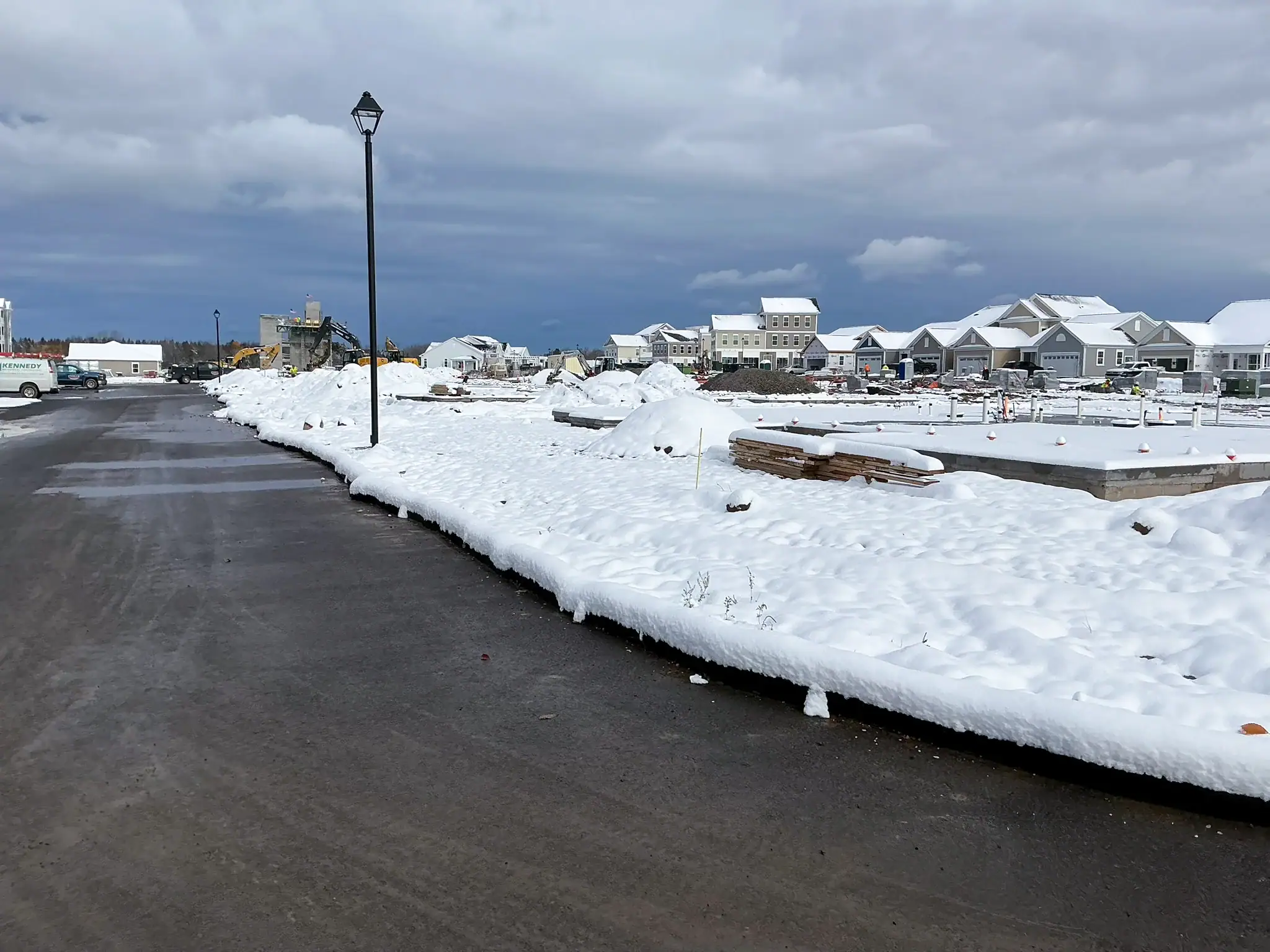 Active residential construction site during winter showing snow-covered building materials, equipment, and partially completed homes under overcast sky at Arbors at Penfield