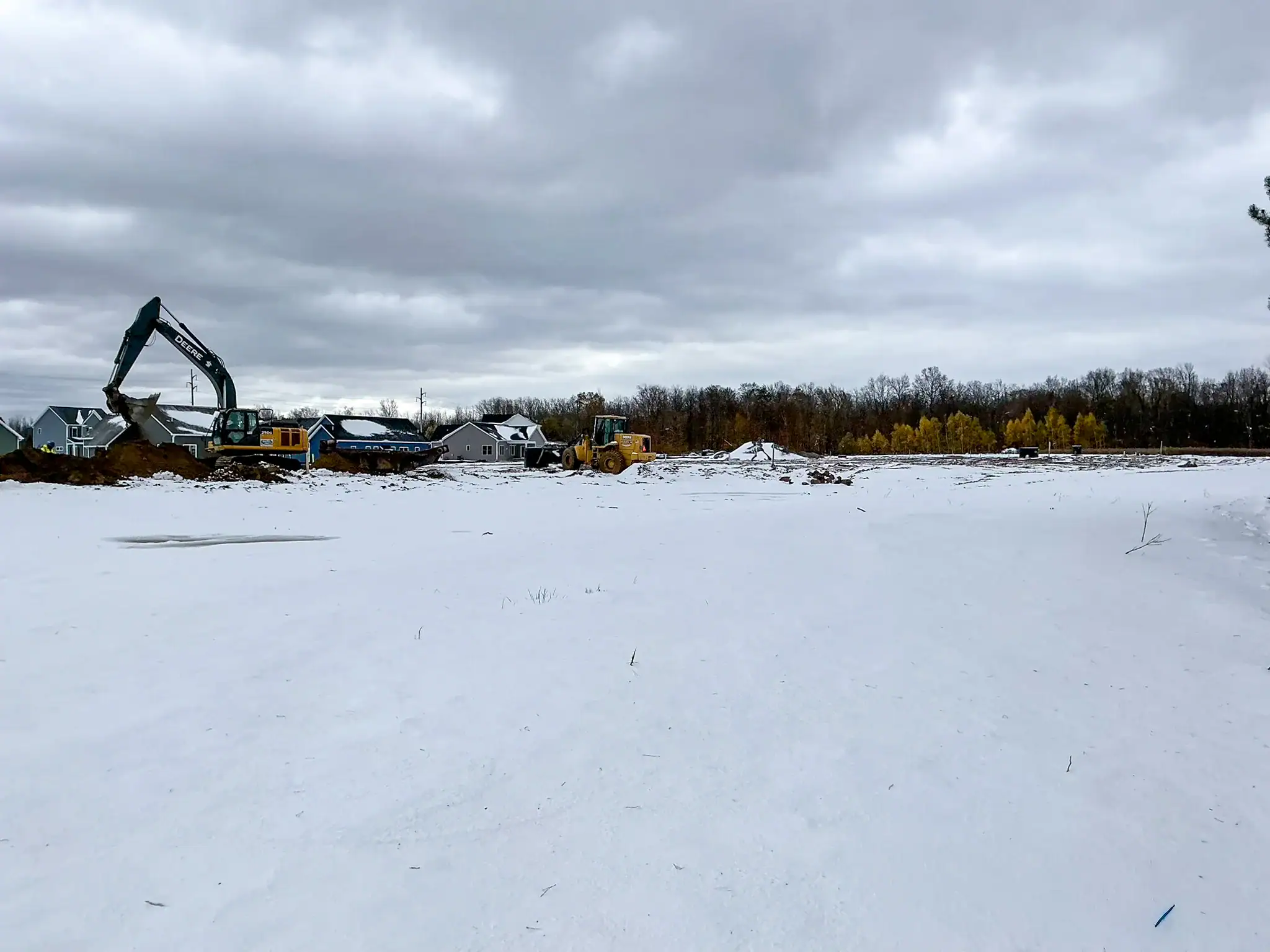 Active winter construction at Arbors at Penfield with excavator and heavy equipment working on snow-covered site with residential homes visible