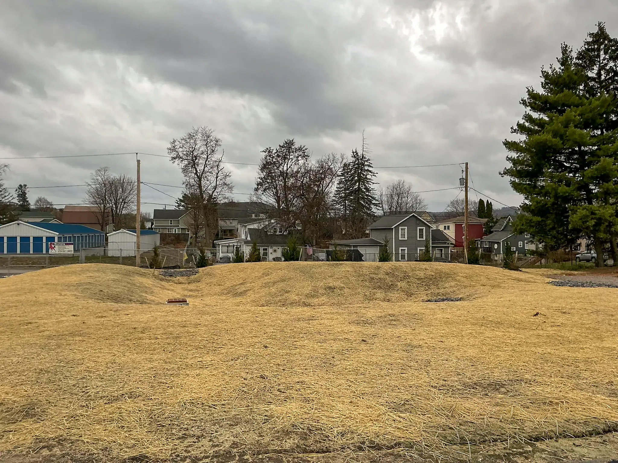 Construction site with straw mulch covering disturbed soil mound in residential neighborhood under overcast sky