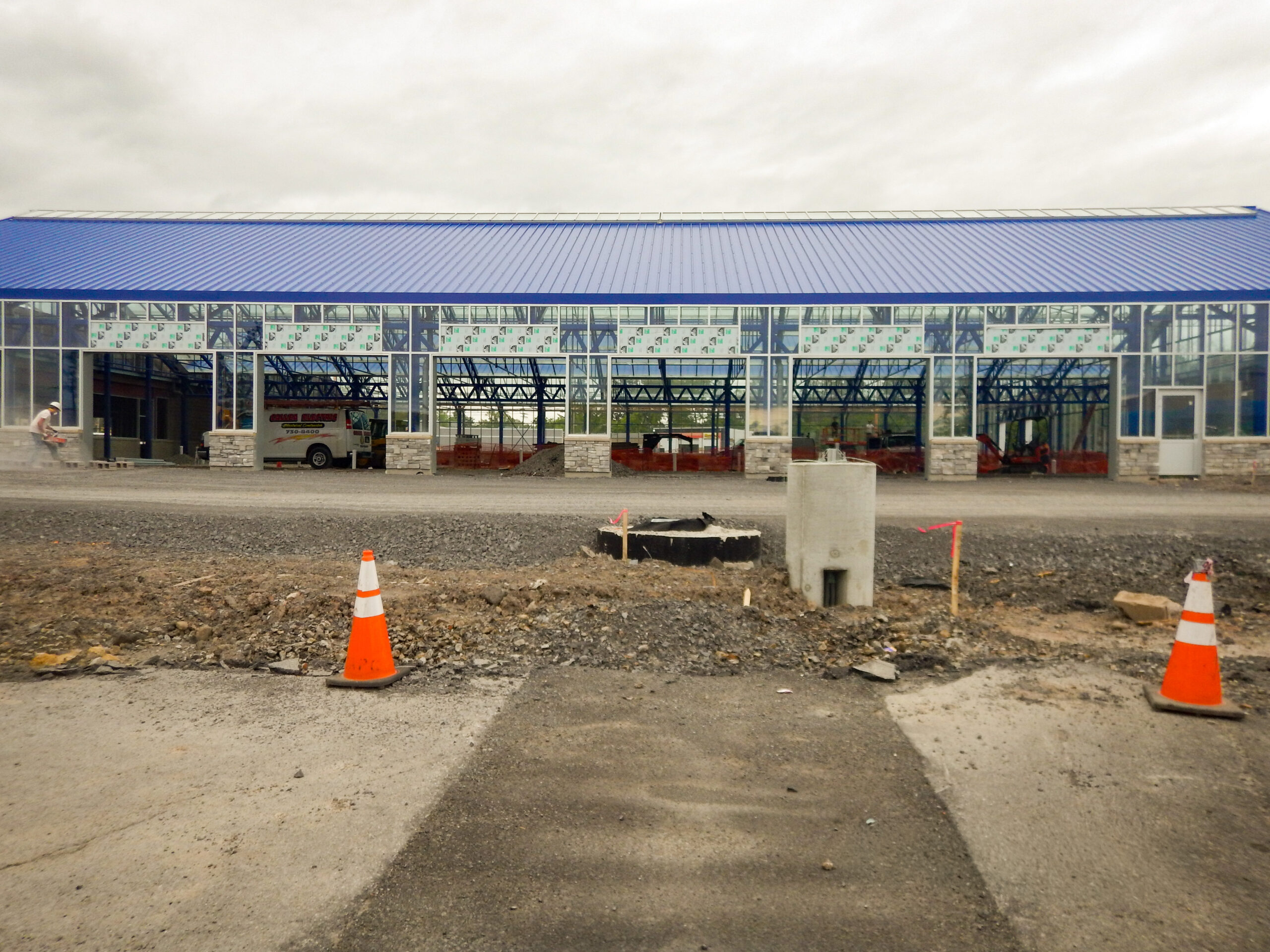 Delta Sonic West Henrietta carwash building under construction showing steel frame, blue metal roof, large glass facade, and site grading with concrete utility structure in foreground.
