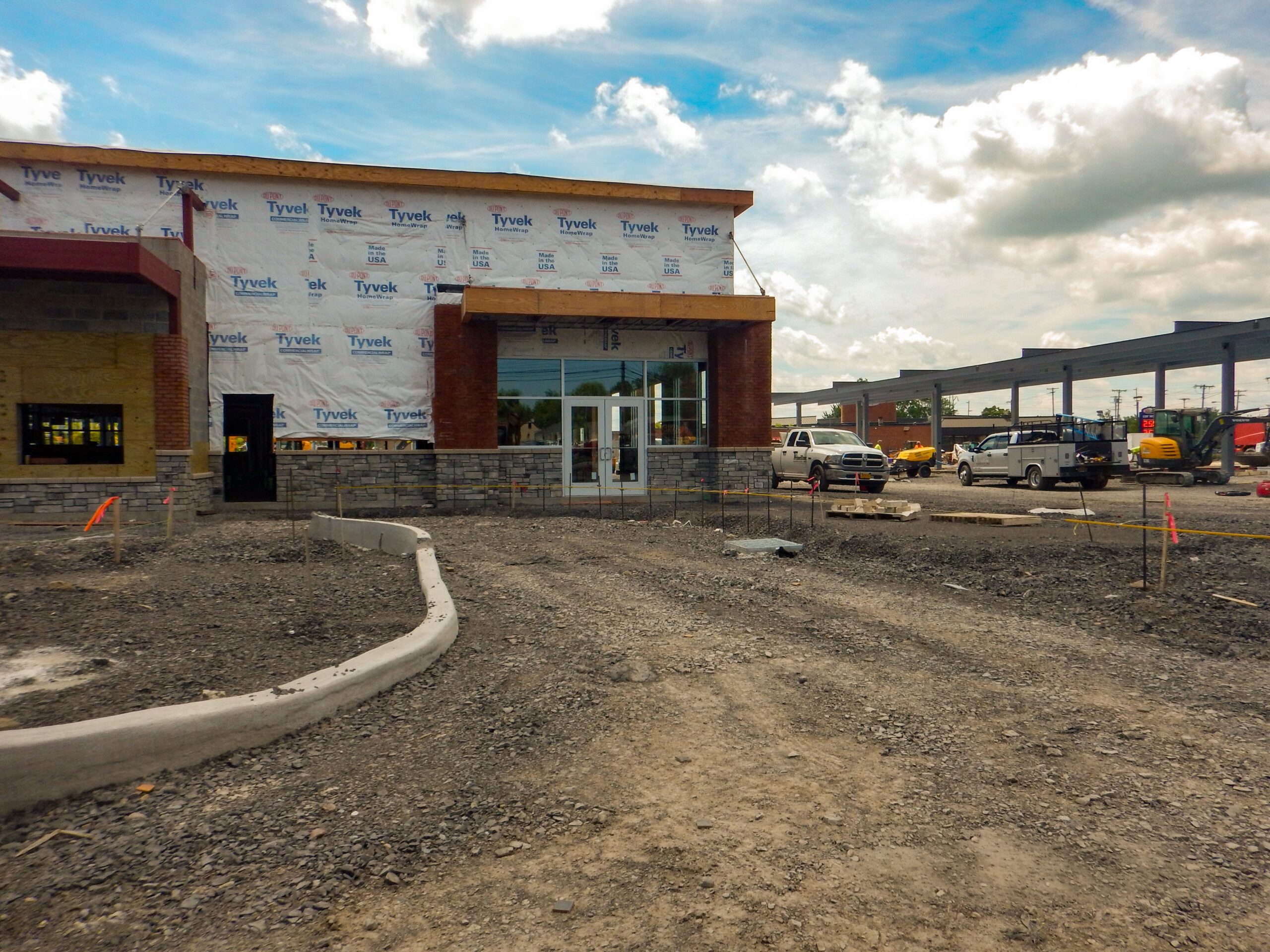 Delta Sonic West Henrietta service building under construction showing brick and stone facade, Tyvek wrapping, concrete site edging, and raw grading during early construction phase.