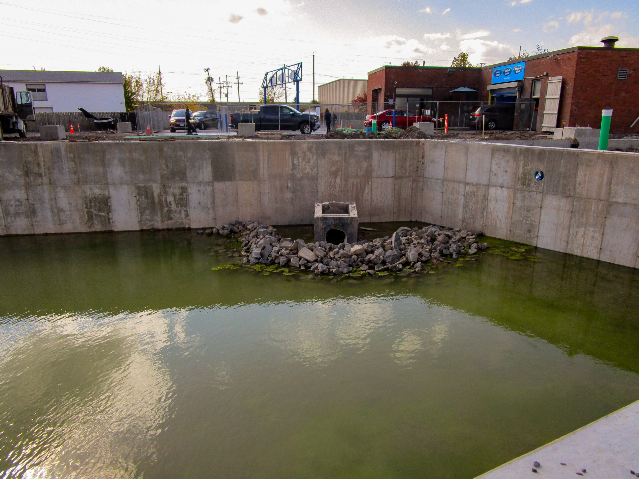 Delta Sonic West Henrietta detention pond showing concrete walls, outlet structure with riprap protection, and facility operations continuing in background during construction.