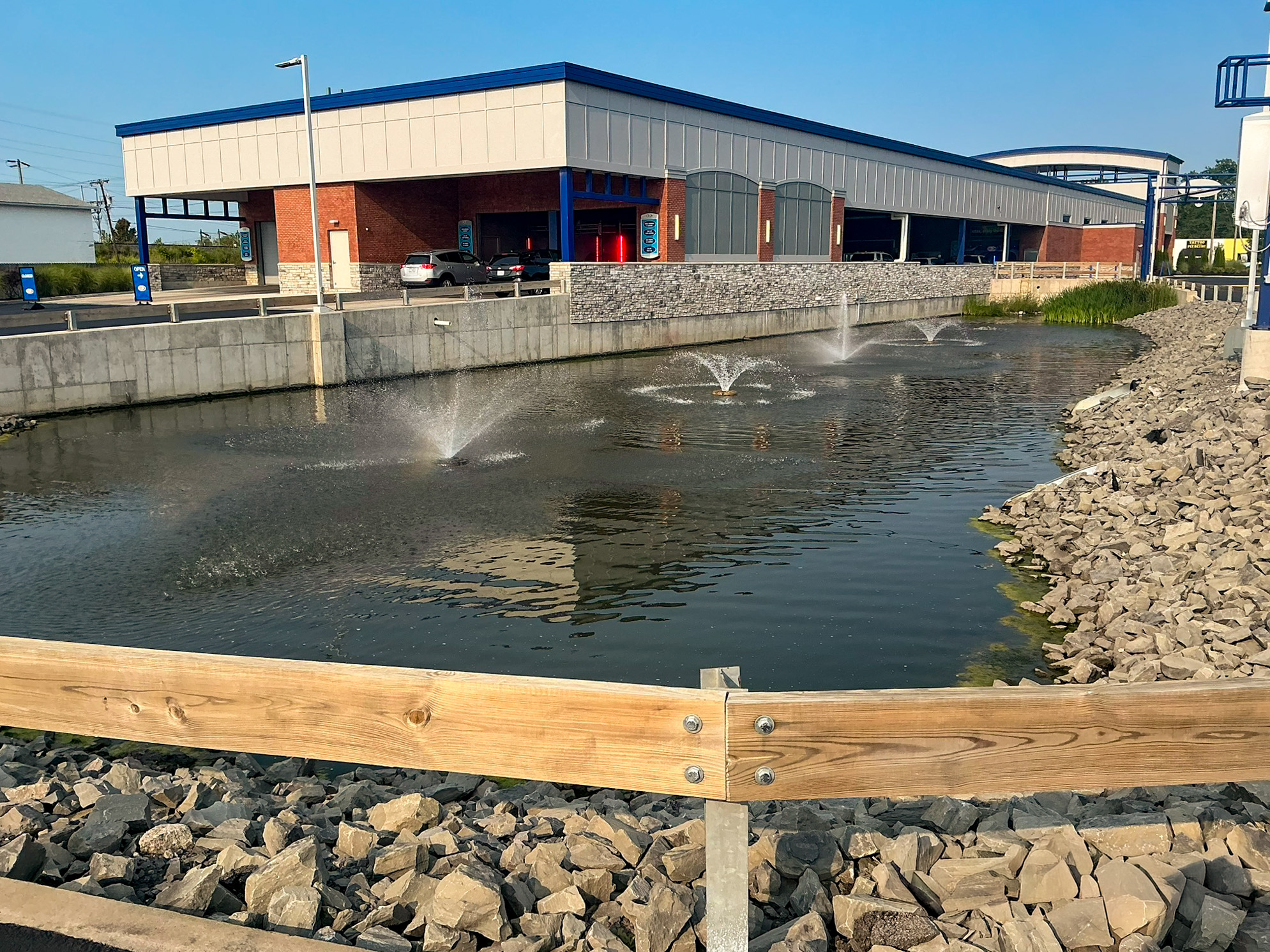 Delta Sonic West Henrietta stormwater detention pond with aerator fountains, riprap bank protection, wooden perimeter fencing, and service buildings visible in background.