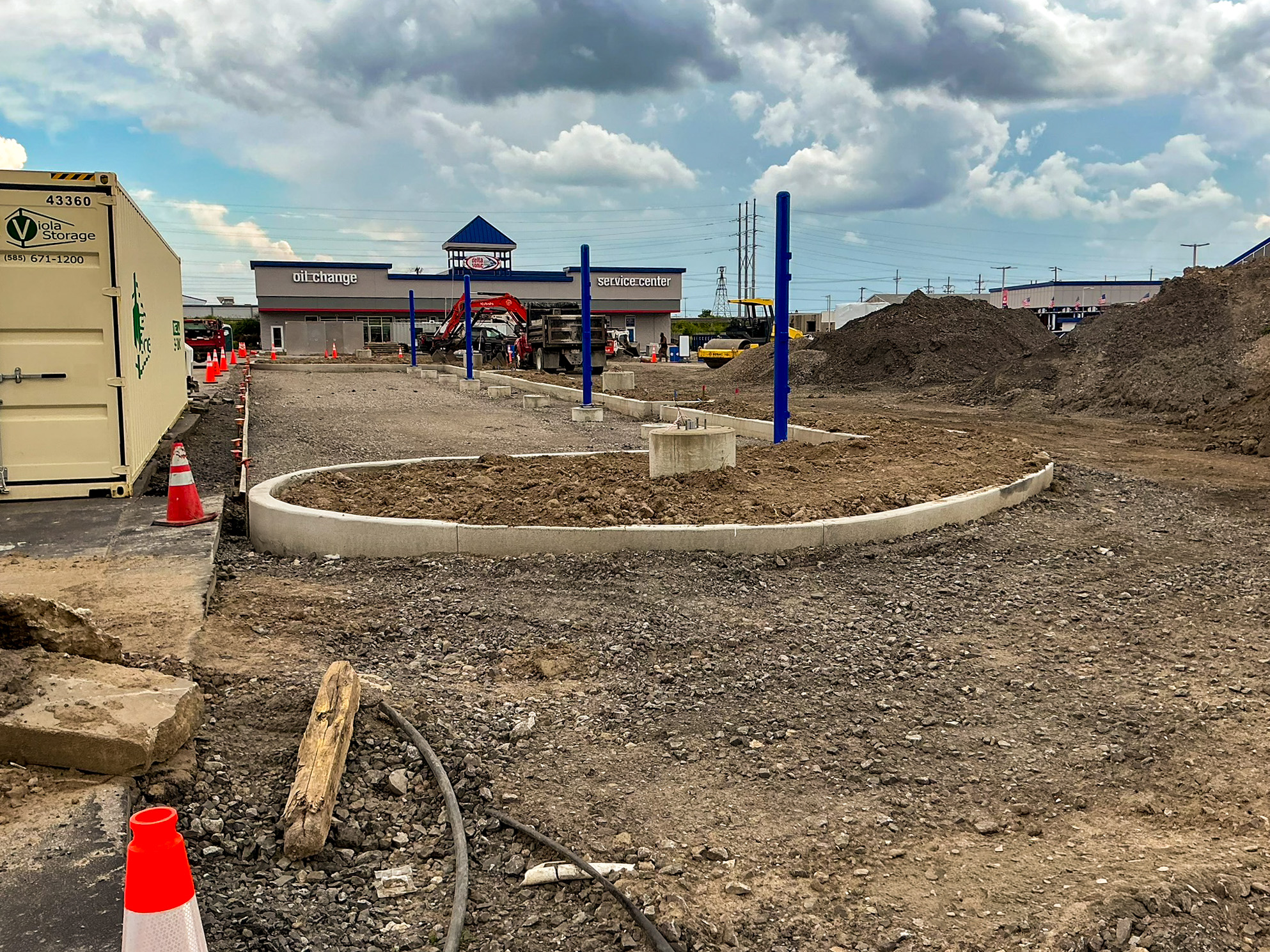 Delta Sonic West Henrietta roundabout construction showing concrete edging structure, lighting pole installation, grading work, and oil change/service buildings in background.